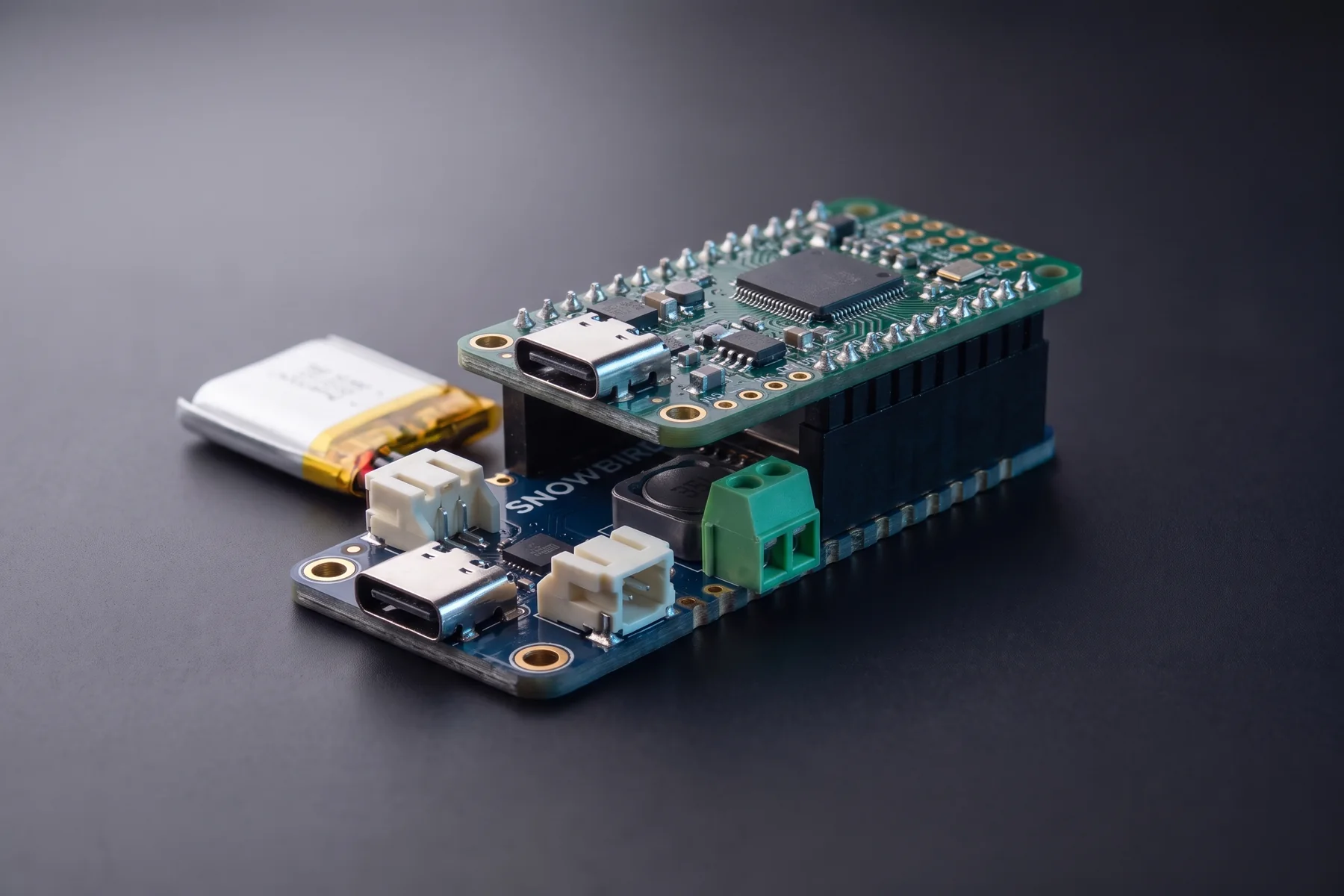A Snowbird Feather 1S with a plain microcontroller Feather stacked on top of it on black stacking headers, a pouch cell plugged into the lower board.